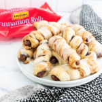 White round plate piled with cinnamon roll pigs in a blanket that are drizzled with icing. Rhodes cinnamon roll package in the background.