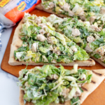 Grilled chicken caesar salad flatbread sliced into triangles on a pizza peel. Package of Rhodes bread dough in the background.