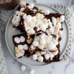 Pile of hot chocolate brownies on a round plate with a mug of hot chocolate to the left of the plate. Waffle weave towel in the forefront.